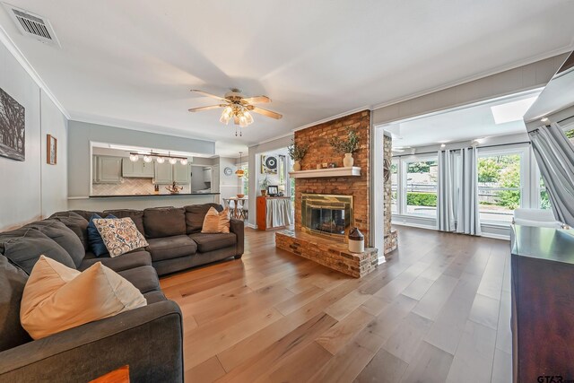 living room featuring wood finished floors, visible vents, a ceiling fan, ornamental molding, and a brick fireplace