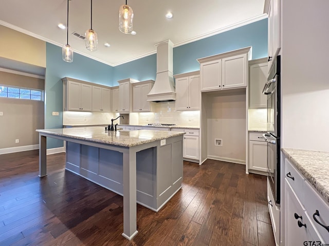 kitchen with a breakfast bar, a kitchen island with sink, hanging light fixtures, light stone countertops, and custom range hood