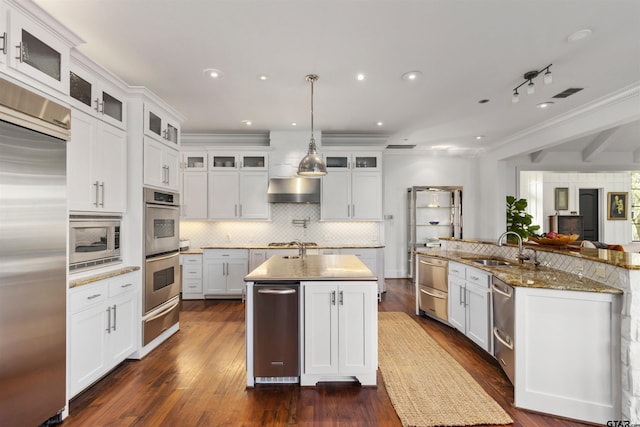 kitchen featuring wall chimney exhaust hood, sink, pendant lighting, built in appliances, and white cabinets