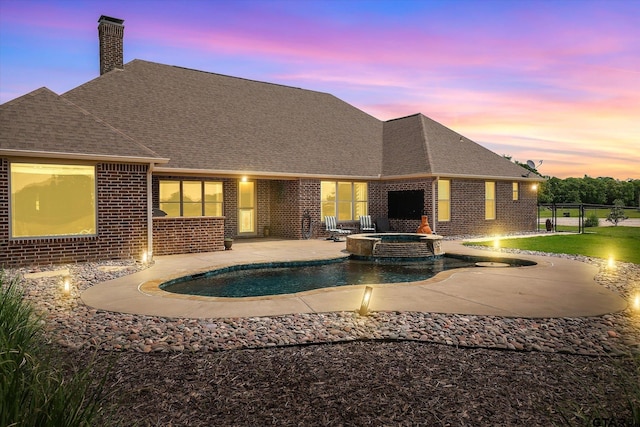 back of house at dusk featuring fence, a shingled roof, brick siding, a chimney, and a patio