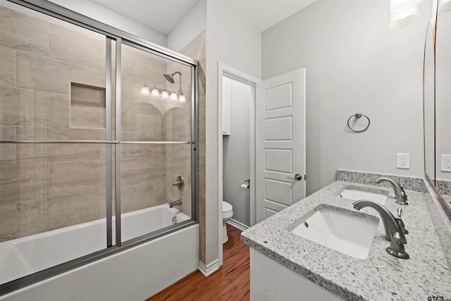 bathroom featuring a sink, double vanity, toilet, wood finished floors, and bath / shower combo with glass door