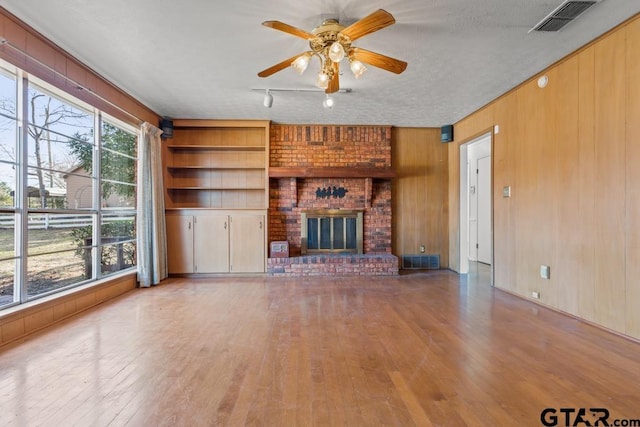 unfurnished living room with a textured ceiling, built in shelves, wooden walls, ceiling fan, and a brick fireplace