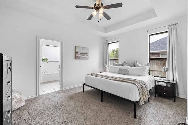 bedroom featuring a ceiling fan, light colored carpet, a raised ceiling, baseboards, and ensuite bathroom