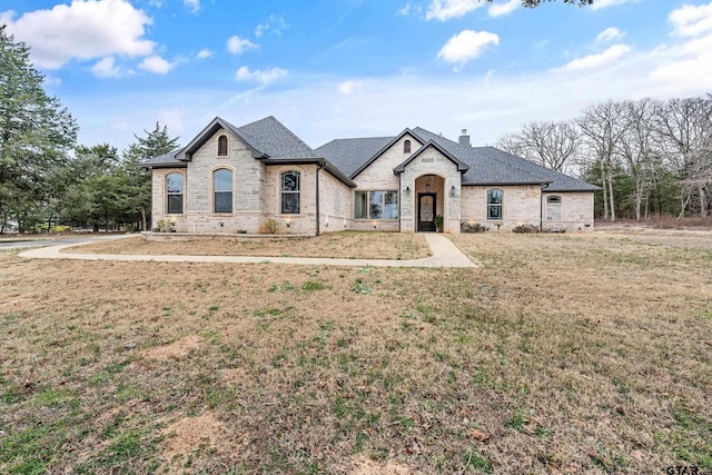 french country style house featuring stone siding, a chimney, a front lawn, and roof with shingles