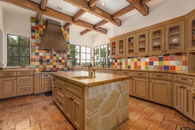 kitchen featuring backsplash, premium range hood, a center island with sink, sink, and beam ceiling