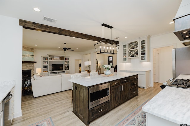 kitchen featuring beamed ceiling, light wood-style flooring, stainless steel appliances, a large fireplace, and white cabinets