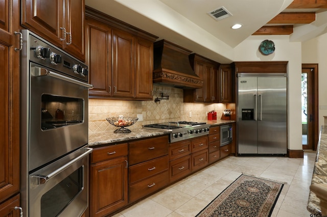 kitchen featuring built in appliances, tasteful backsplash, light tile patterned flooring, light stone countertops, and premium range hood