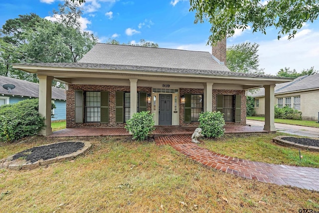 view of front of property featuring a front yard, covered porch, a shingled roof, a chimney, and brick siding