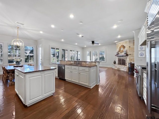 kitchen featuring hanging light fixtures, stainless steel appliances, white cabinets, a center island with sink, and a stone fireplace