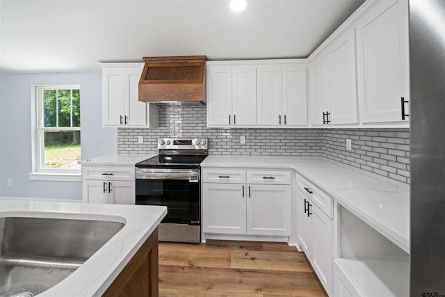 kitchen with light stone counters, custom exhaust hood, stainless steel electric stove, white cabinetry, and light hardwood / wood-style flooring