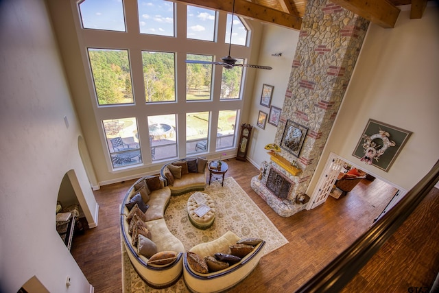 living room with hardwood / wood-style floors, a high ceiling, a stone fireplace, ceiling fan, and beam ceiling