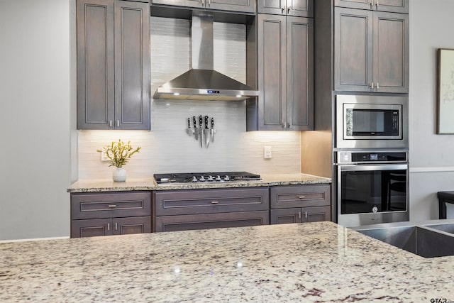 kitchen featuring tasteful backsplash, light stone counters, wall chimney range hood, and appliances with stainless steel finishes