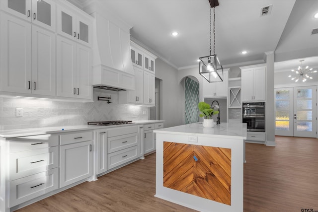 kitchen featuring custom exhaust hood, a kitchen island with sink, white cabinets, double oven, and decorative light fixtures