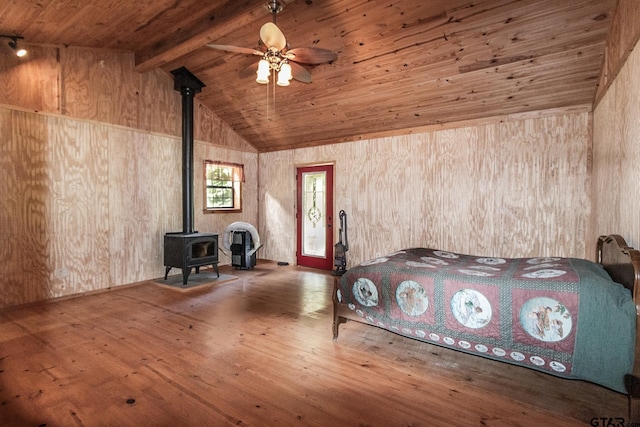 unfurnished bedroom featuring a wood stove, ceiling fan, vaulted ceiling with beams, hardwood / wood-style floors, and wooden walls