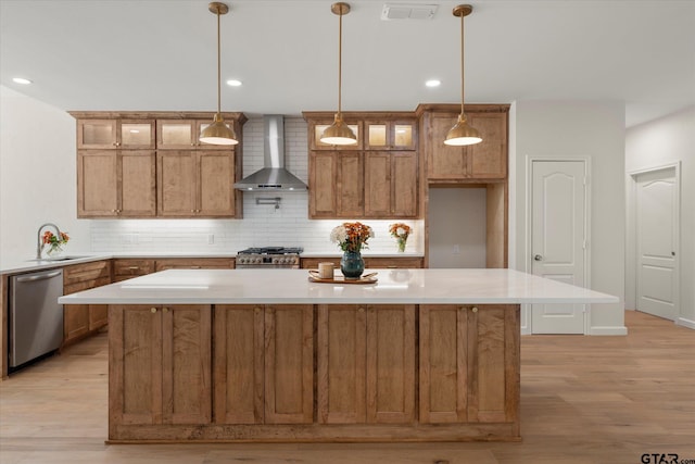 kitchen with wall chimney range hood, appliances with stainless steel finishes, decorative light fixtures, light wood-type flooring, and a center island