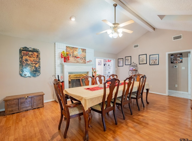 dining room featuring a textured ceiling, vaulted ceiling with beams, light wood-style flooring, visible vents, and a brick fireplace