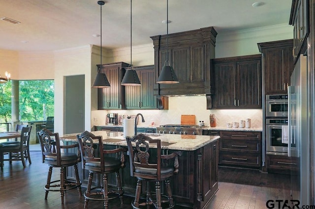 kitchen featuring dark wood-type flooring, hanging light fixtures, a center island with sink, light stone countertops, and decorative backsplash