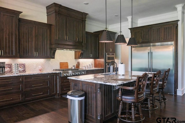 kitchen with appliances with stainless steel finishes, hanging light fixtures, light stone counters, tasteful backsplash, and an island with sink