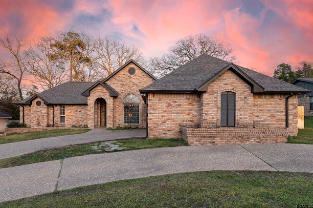 french country home with driveway, brick siding, roof with shingles, and a front yard