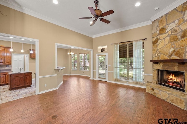 unfurnished living room featuring a sink, a stone fireplace, ceiling fan with notable chandelier, light wood-style floors, and ornamental molding