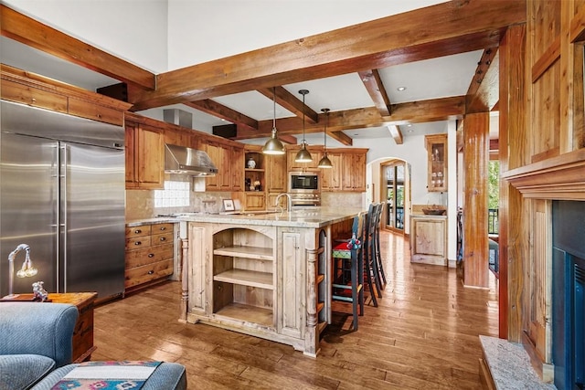 kitchen featuring wall chimney exhaust hood, tasteful backsplash, built in appliances, a spacious island, and decorative light fixtures