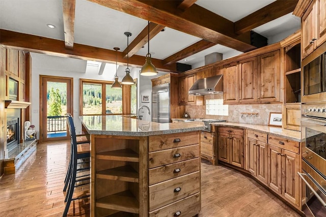 kitchen featuring tasteful backsplash, wall chimney exhaust hood, stainless steel appliances, decorative light fixtures, and an island with sink