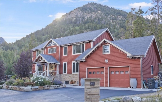 view of front of house with driveway, stone siding, a mountain view, a wooded view, and a garage