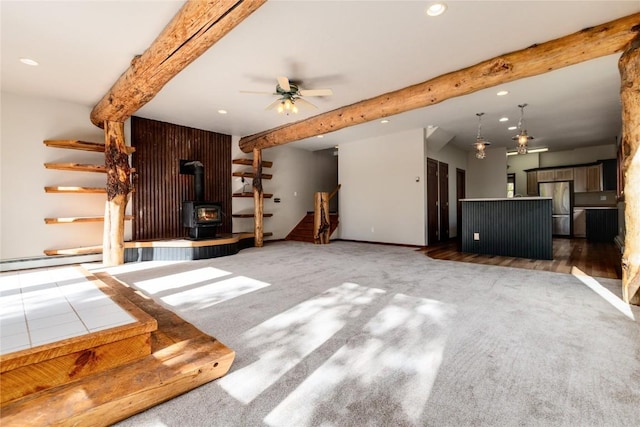 unfurnished living room with a wood stove, dark colored carpet, wooden walls, ceiling fan, and beam ceiling