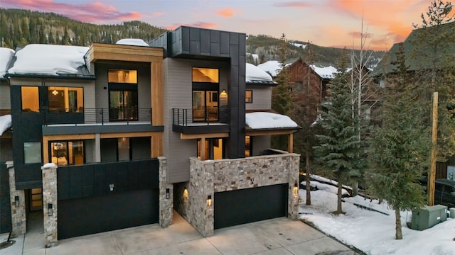 modern home featuring driveway, a standing seam roof, stone siding, metal roof, and a balcony