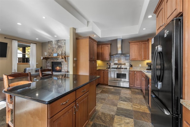 kitchen with a center island, black appliances, a stone fireplace, dark stone counters, and wall chimney exhaust hood