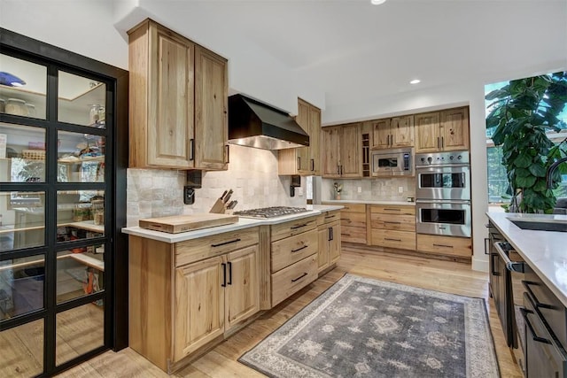 kitchen featuring decorative backsplash, stainless steel appliances, light countertops, light wood-type flooring, and wall chimney range hood