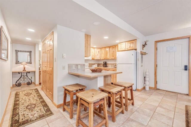 kitchen featuring kitchen peninsula, light brown cabinets, white fridge, a breakfast bar area, and light tile patterned flooring