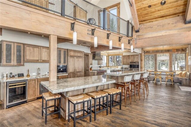 kitchen featuring high vaulted ceiling, wall chimney range hood, hanging light fixtures, wine cooler, and light stone counters