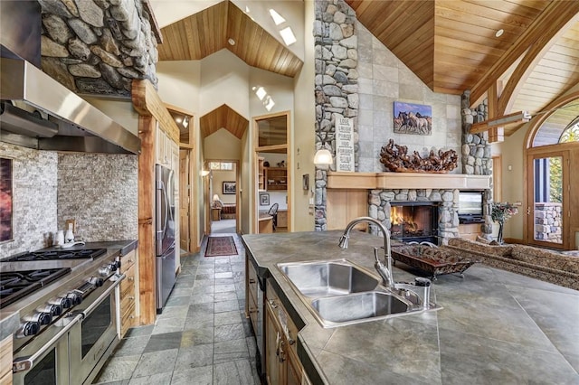 kitchen featuring stainless steel appliances, wall chimney range hood, sink, high vaulted ceiling, and a stone fireplace