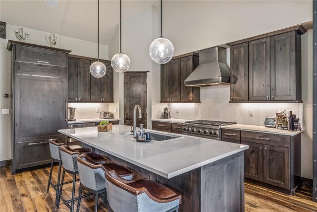 kitchen with wall chimney exhaust hood, dark brown cabinetry, a kitchen island with sink, sink, and hanging light fixtures