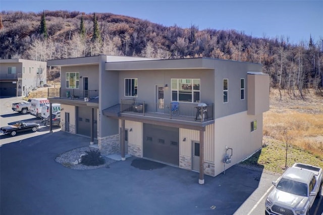 view of front of house featuring aphalt driveway, stone siding, a view of trees, a garage, and a balcony