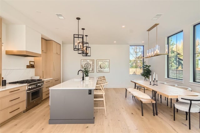 kitchen featuring hanging light fixtures, high end stove, light brown cabinets, custom exhaust hood, and a kitchen island with sink