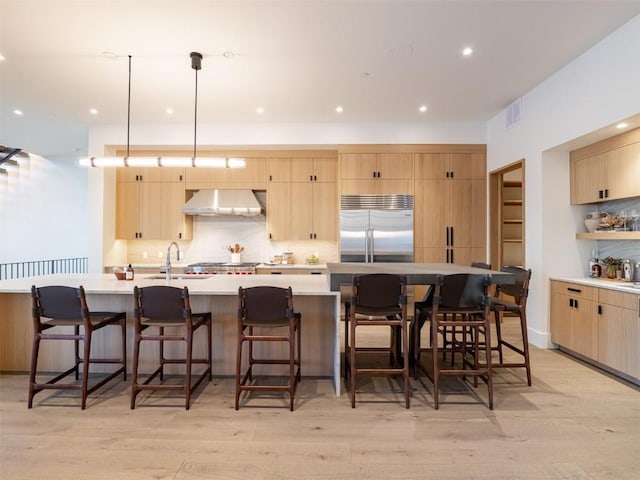 kitchen featuring wall chimney exhaust hood, stainless steel appliances, an island with sink, light hardwood / wood-style floors, and light brown cabinetry