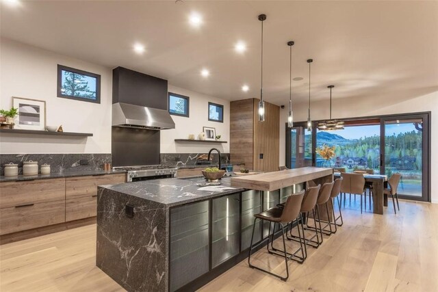 kitchen with hanging light fixtures, wall chimney range hood, a mountain view, light hardwood / wood-style floors, and a kitchen island with sink