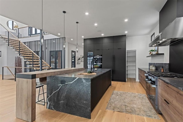 kitchen featuring a spacious island, light hardwood / wood-style flooring, wall chimney exhaust hood, high end stove, and decorative light fixtures