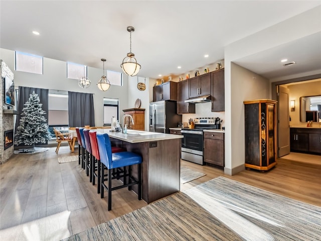 kitchen featuring appliances with stainless steel finishes, hanging light fixtures, light wood-type flooring, a center island with sink, and dark brown cabinets