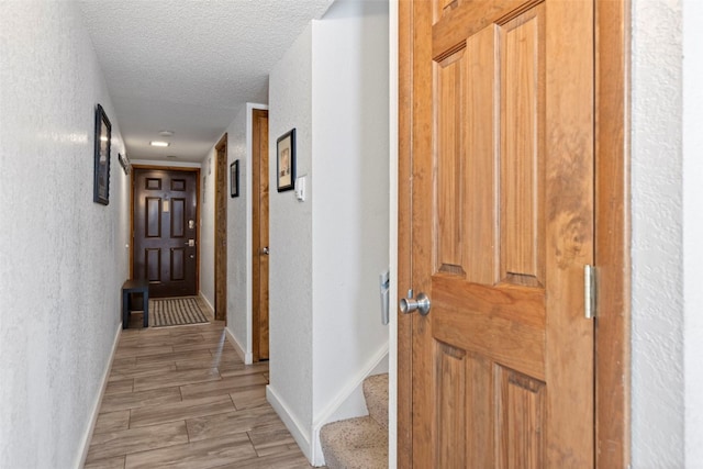 corridor with light wood finished floors, a textured wall, baseboards, and a textured ceiling