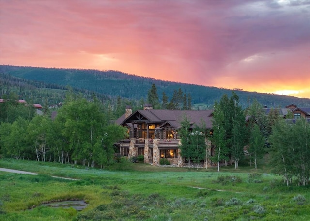 back of house at dusk featuring a balcony, a forest view, stone siding, a chimney, and a mountain view