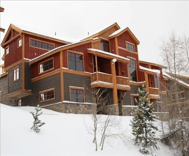 snow covered rear of property featuring stone siding, a balcony, and board and batten siding