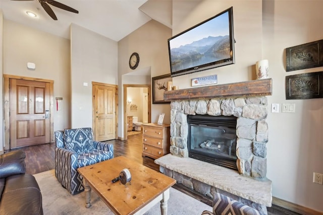 living area featuring a ceiling fan, high vaulted ceiling, a stone fireplace, and wood finished floors