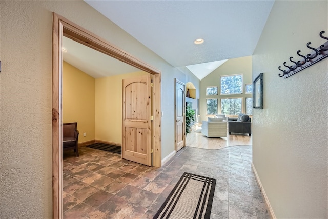 hall featuring lofted ceiling, a textured wall, stone finish floor, and baseboards