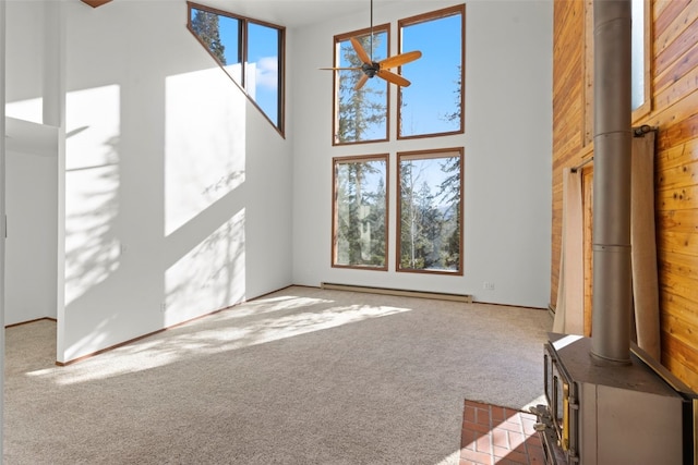unfurnished living room with a wood stove, a baseboard radiator, ceiling fan, a towering ceiling, and carpet flooring