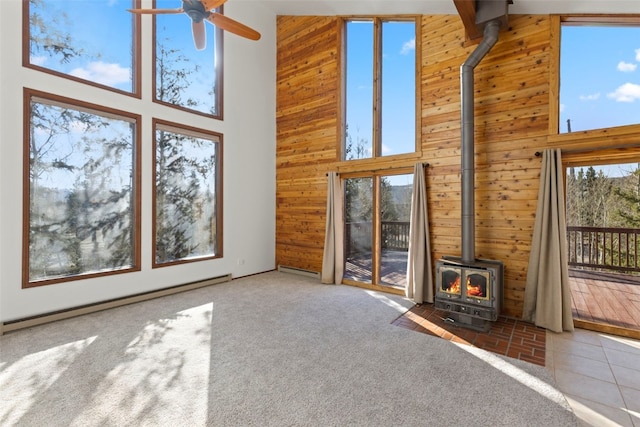 unfurnished living room featuring a wood stove, baseboard heating, wooden walls, a high ceiling, and a ceiling fan