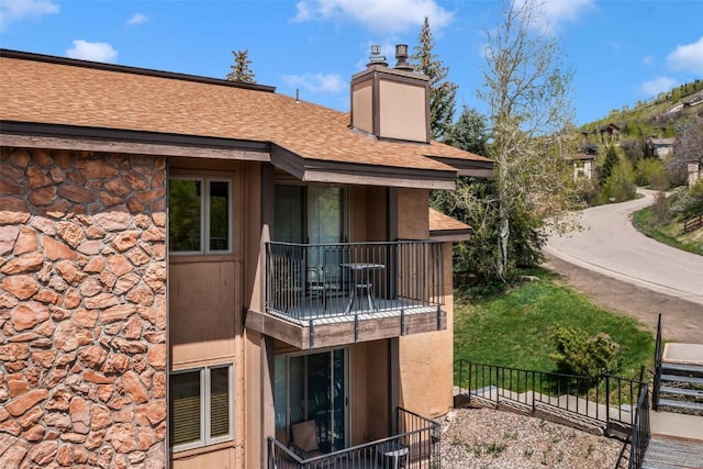 back of house with a balcony, roof with shingles, a chimney, fence, and stone siding