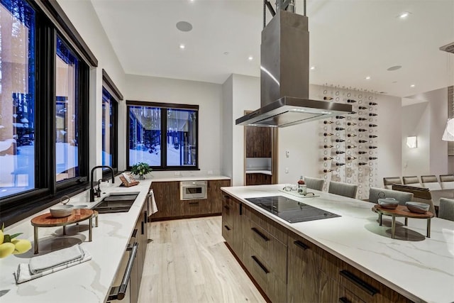 kitchen featuring black electric stovetop, light wood-type flooring, light stone counters, island range hood, and sink